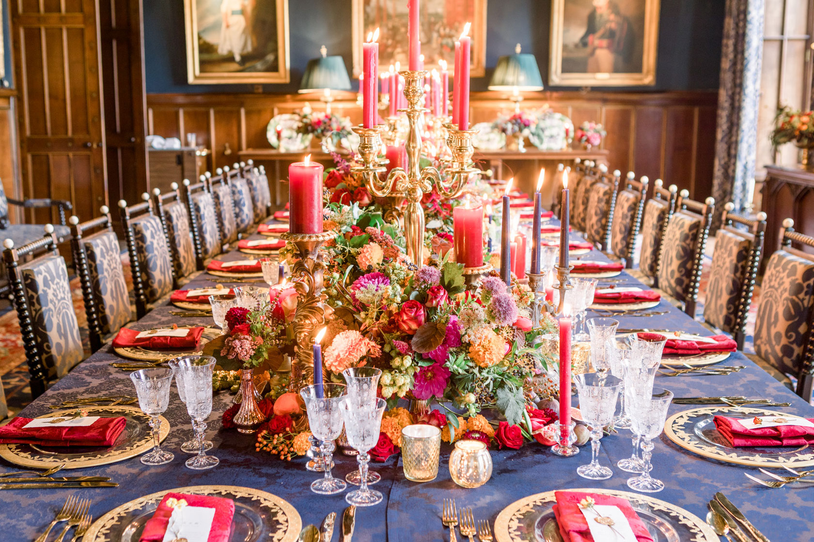 a long table regal inspired wedding brekfast table in a castle with navy linen, gold charger plates and cutlery, gold candelabras with red candles