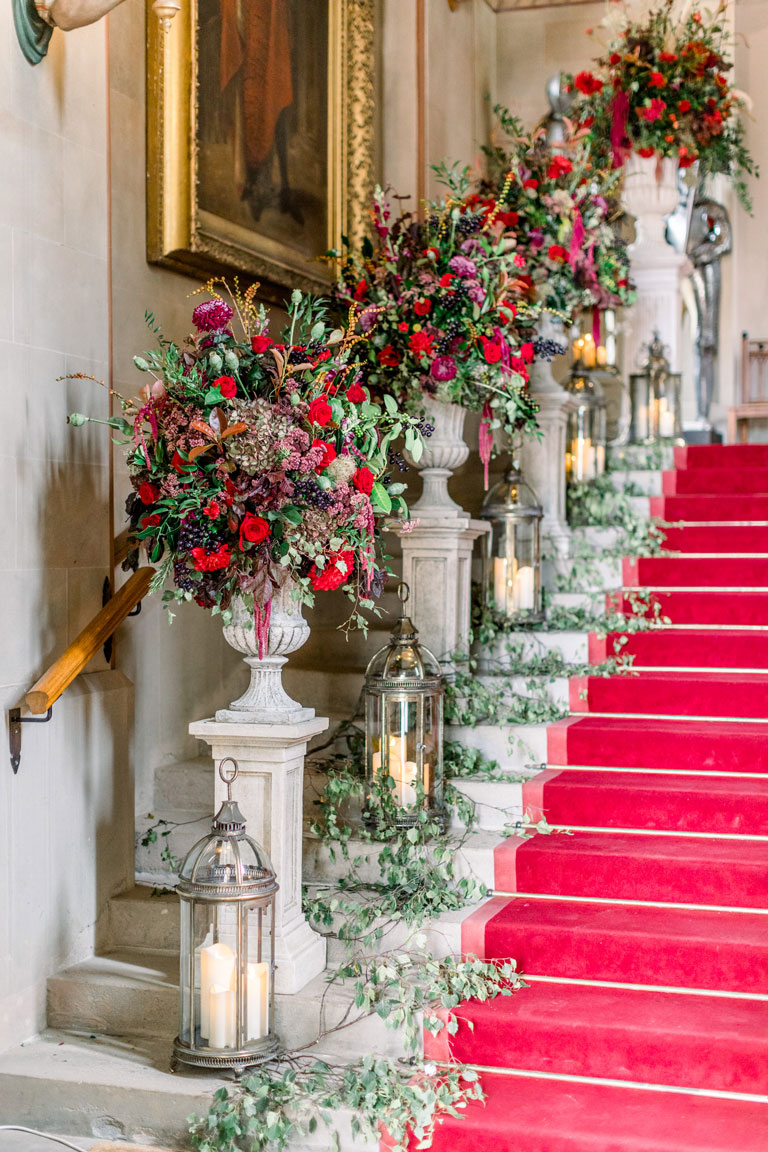 a castle staircase with a red carpet and plynths of regal flowers on each step